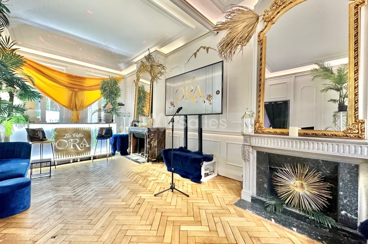Salle élégante avec parquet en chevrons, miroirs dorés, décoration végétale et écran affichant ’ORA’.