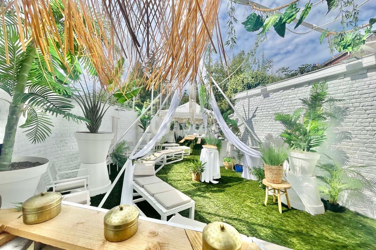 Terrasse extérieure aménagée avec chaises longues, voilages blancs, plantes tropicales et pelouse artificielle.