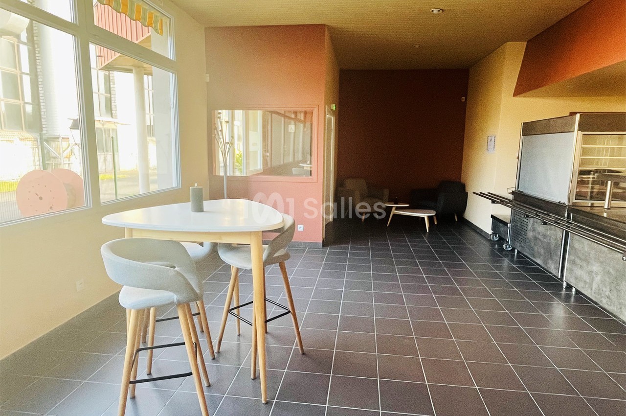 Salle lumineuse avec table haute, chaises, coin salon et grandes fenêtres donnant sur l'extérieur.
