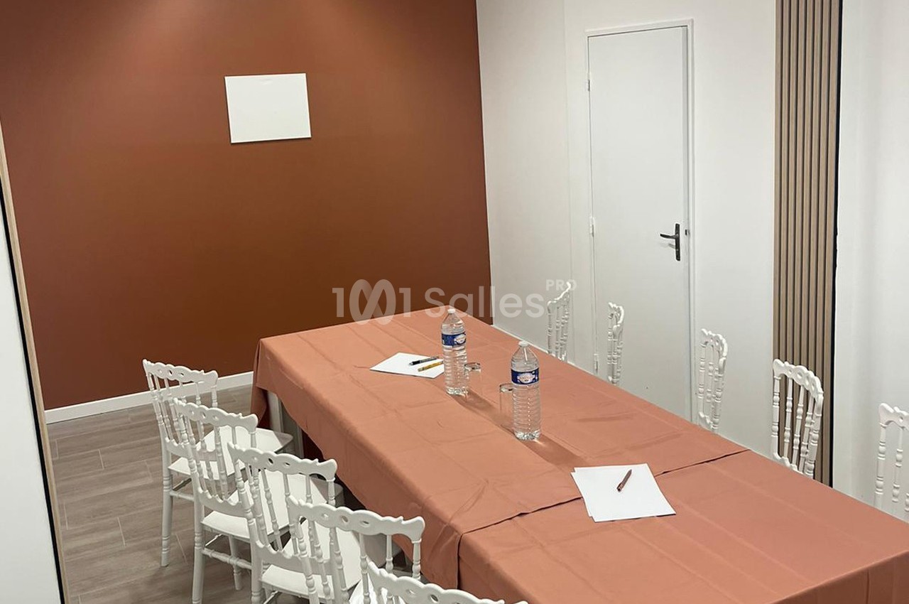 Salle de réunion avec table rectangulaire couverte d'une nappe orange, chaises blanches et fournitures de bureau.