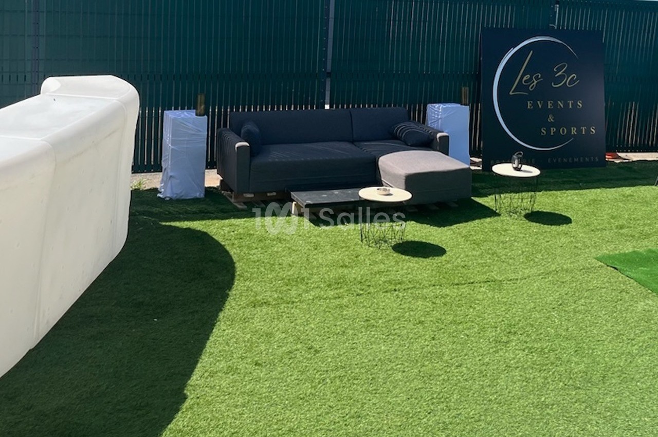 Canapé gris avec table basse sur pelouse synthétique, près d'une enseigne ’Les 3C Events & Sports’.