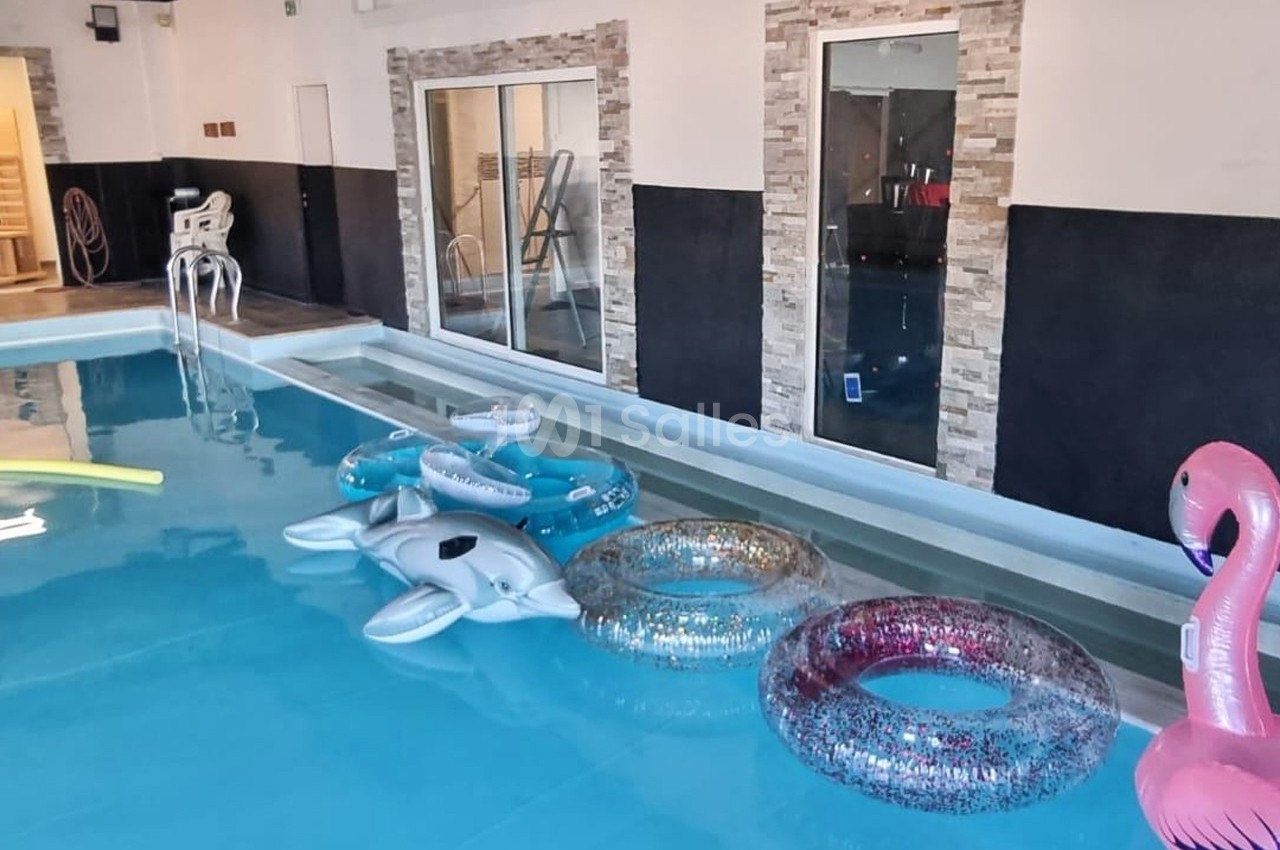 Piscine intérieure avec des bouées colorées flottant à la surface, entourée de murs en pierre et de baies vitrées.