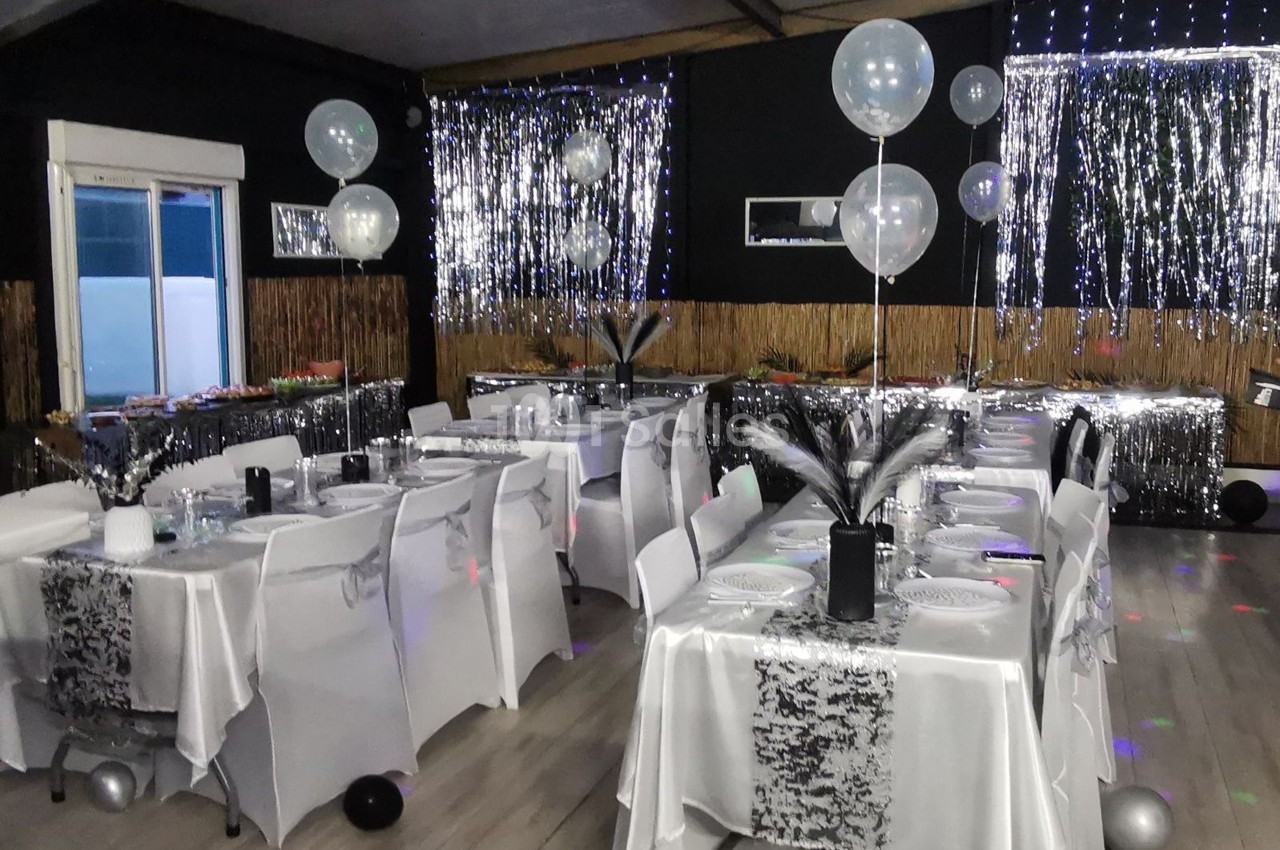 Salle décorée pour une fête avec tables dressées, nappes blanches, ballons transparents et guirlandes argentées.