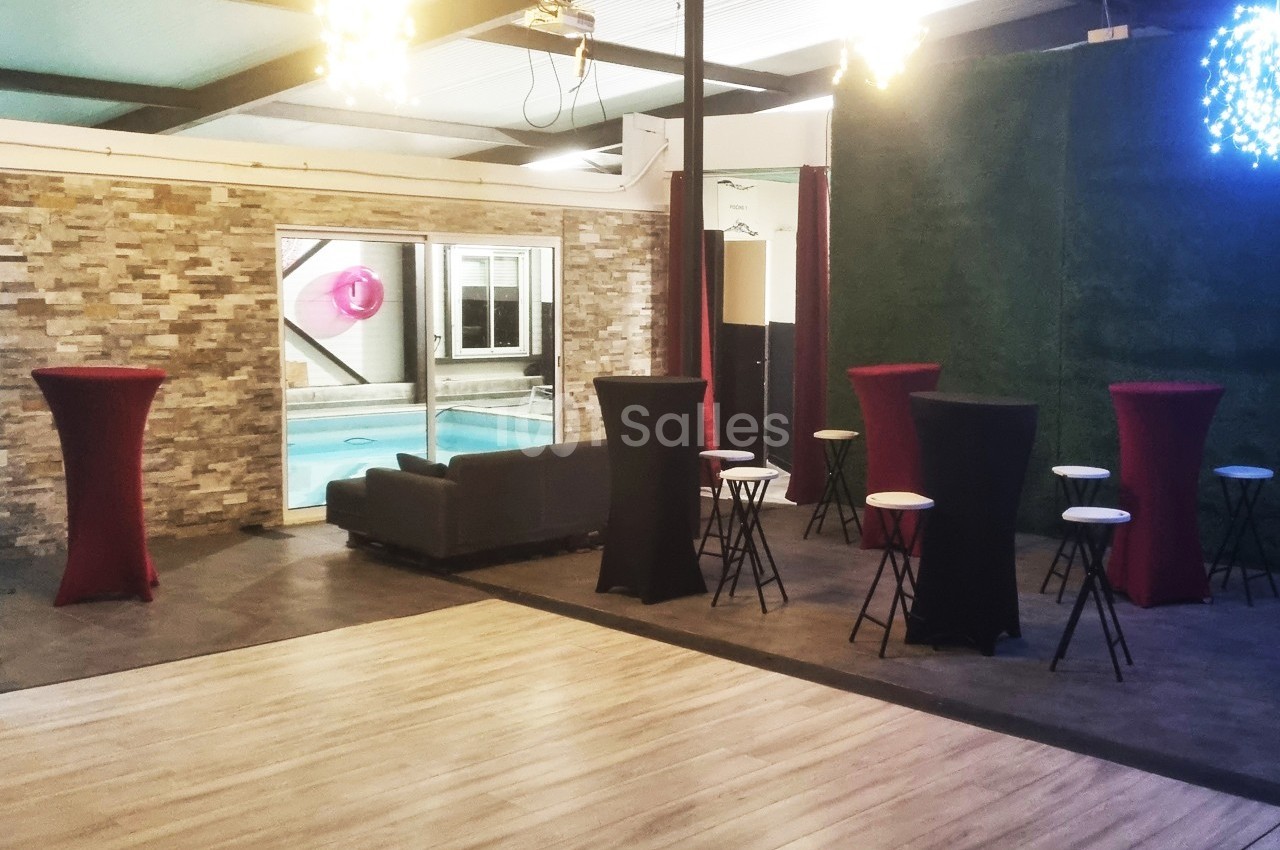Espace intérieur avec tables hautes, chaises, canapé et vue sur une piscine à travers une baie vitrée.