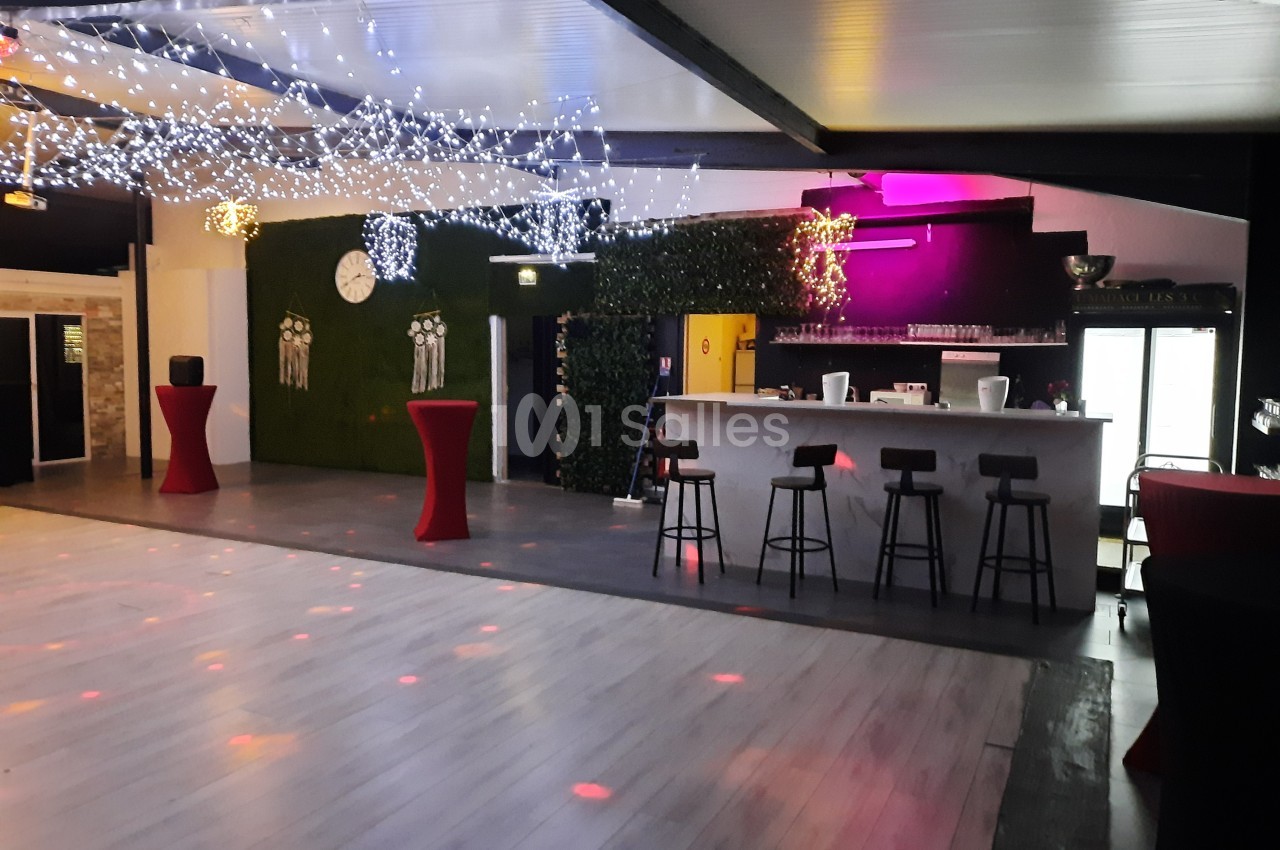 Salle de réception avec piste de danse, bar équipé de tabourets, tables hautes et décorations lumineuses au plafond.