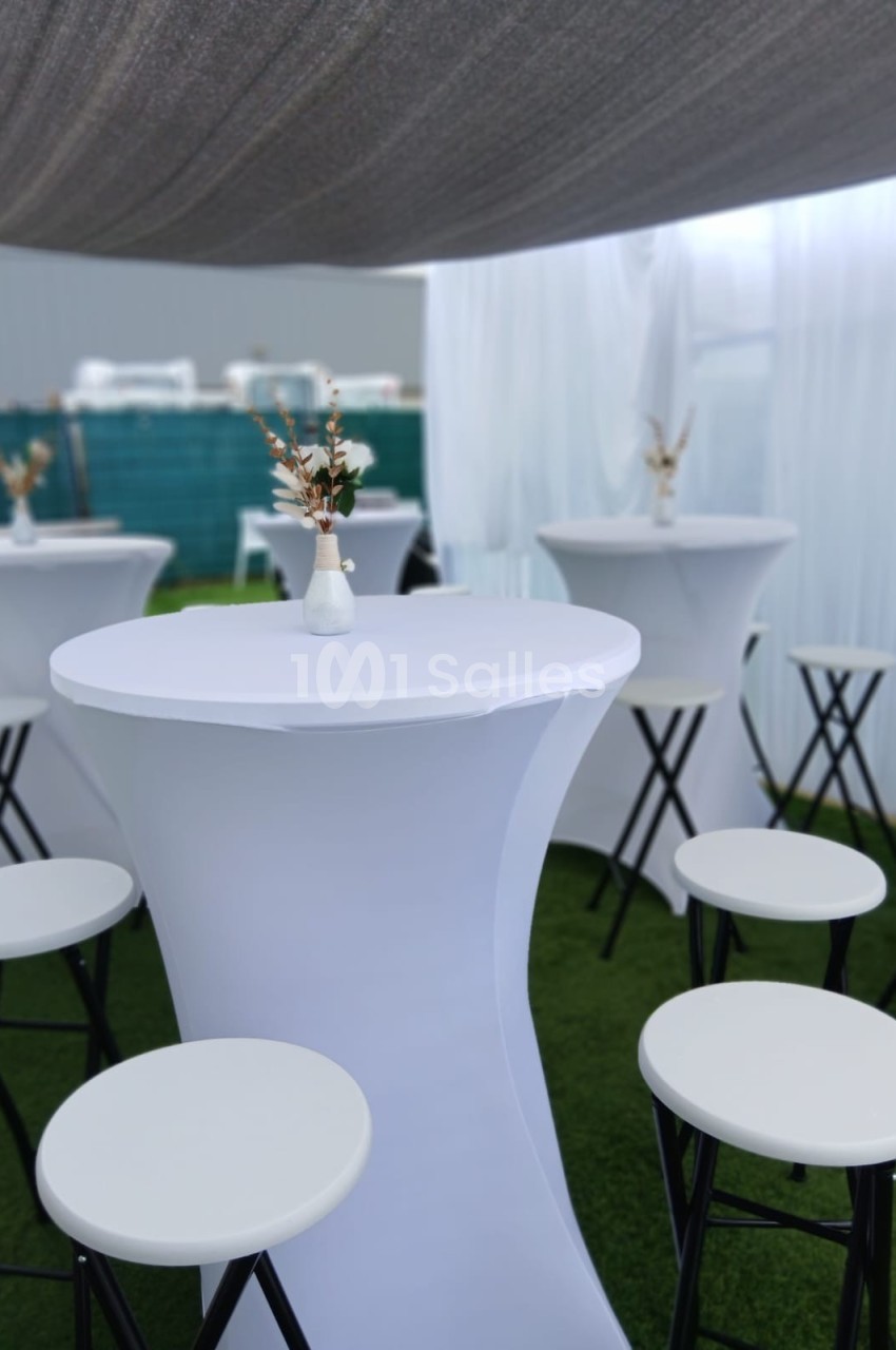 Tables hautes blanches avec tabourets assortis, décorées de petits vases fleuris, dans un espace extérieur couvert.