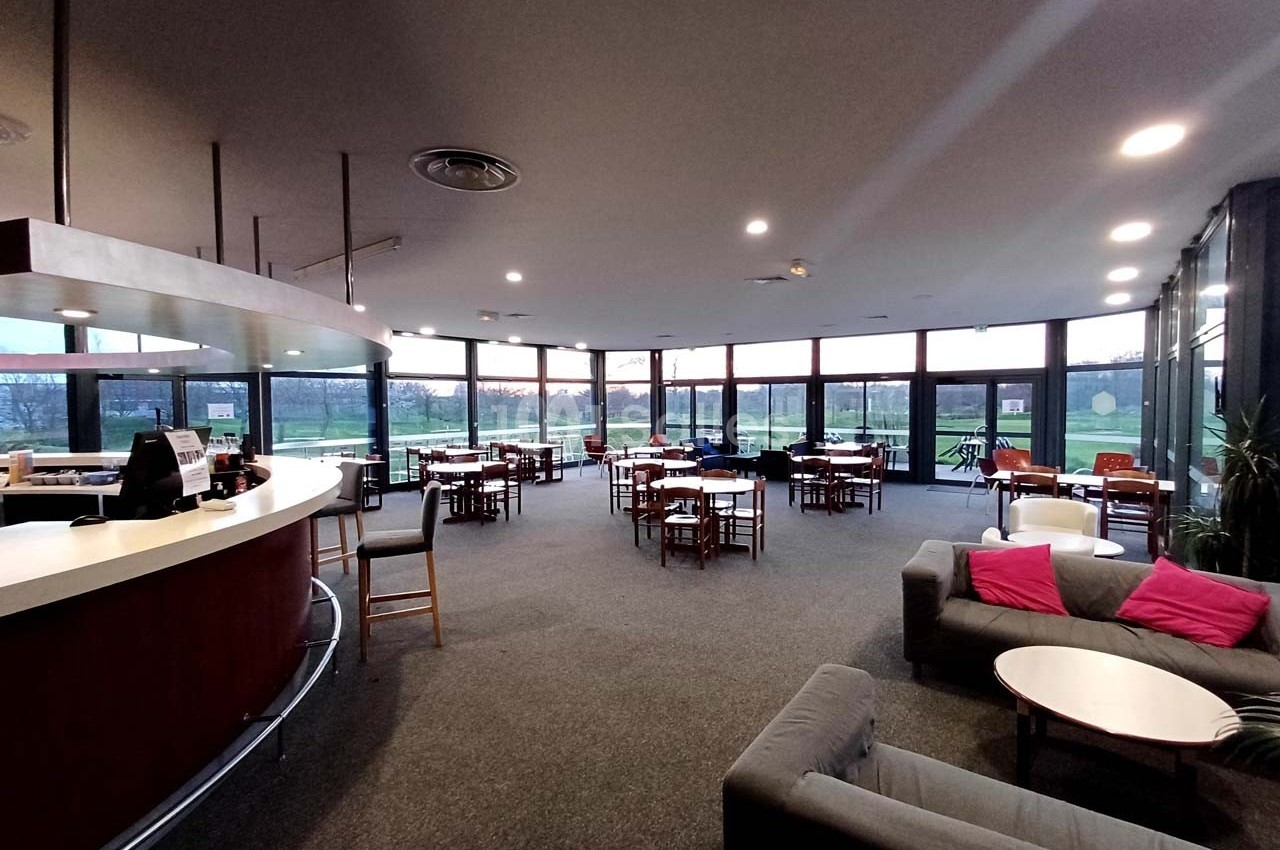 Salle lumineuse avec de grandes baies vitrées, des tables et des chaises disposées autour d'un bar arrondi.