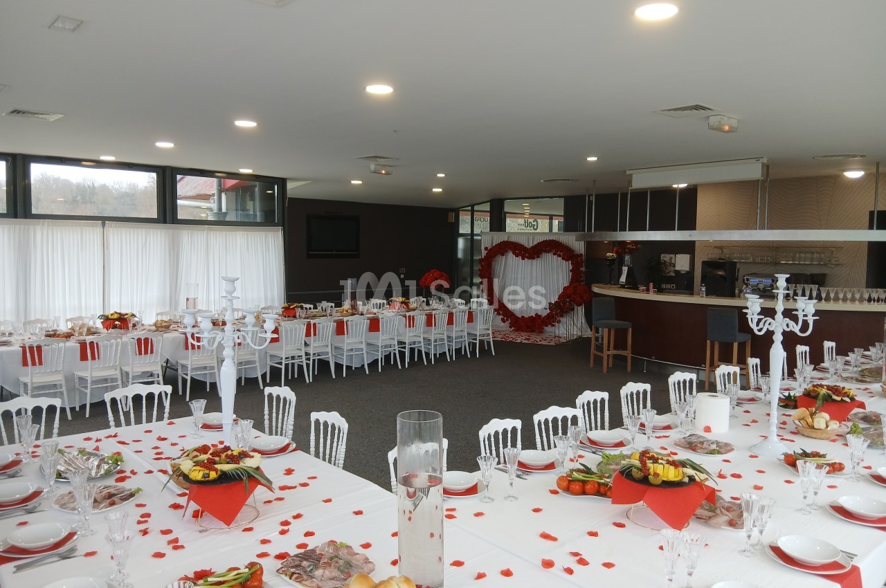 Salle de réception décorée pour un événement, avec tables dressées, pétales rouges et un grand cœur en fond.