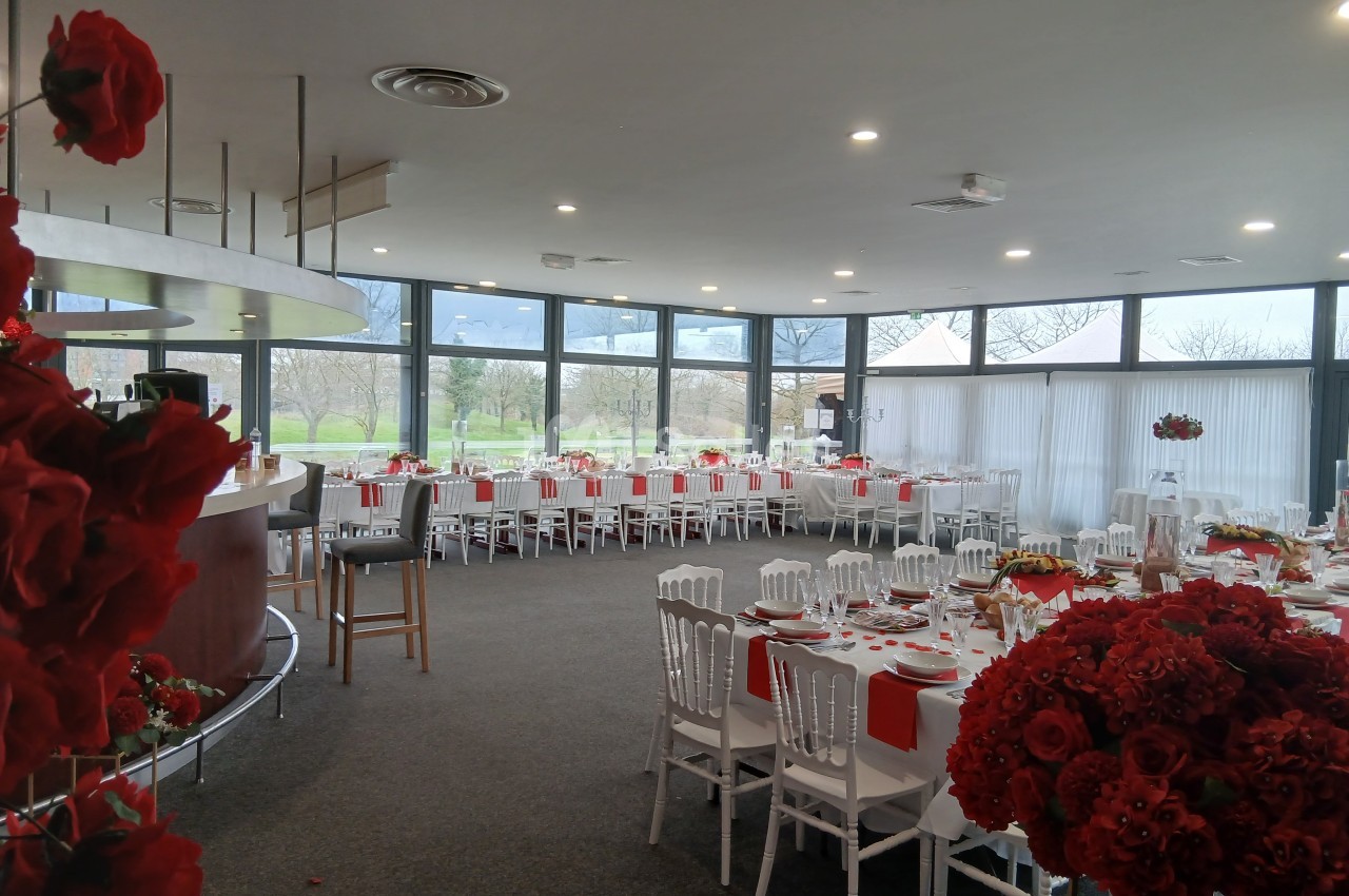 Salle de réception lumineuse avec tables décorées en rouge et blanc, grandes baies vitrées donnant sur un extérieur…
