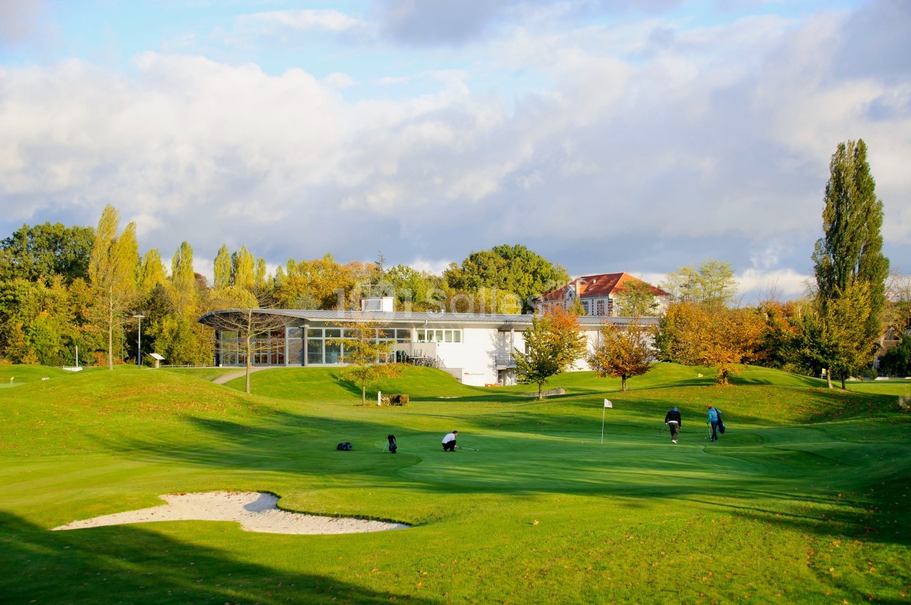 Vue d'un terrain de golf avec des joueurs sur le green, entouré d'arbres et d'un bâtiment en arrière-plan.