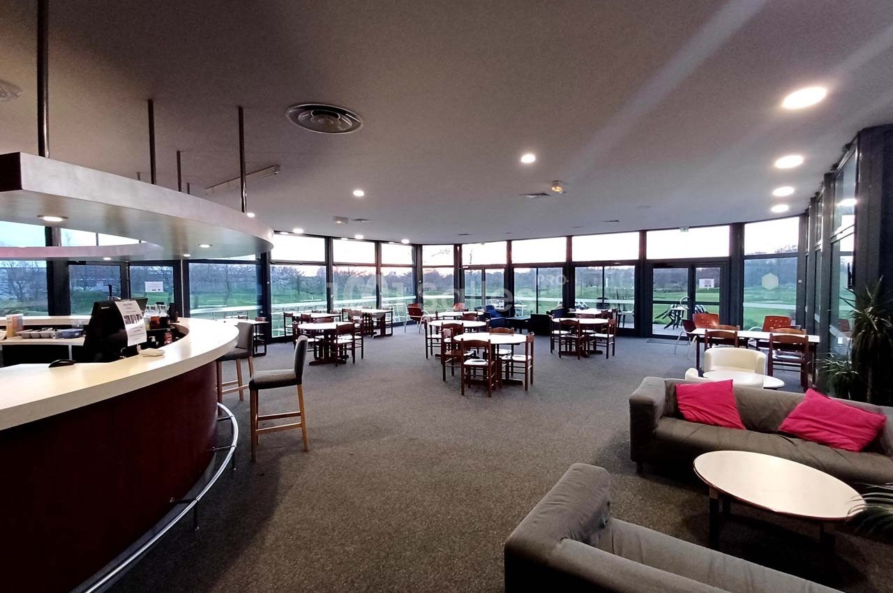 Salle lumineuse avec de grandes baies vitrées, des tables et des chaises disposées autour d'un bar arrondi.