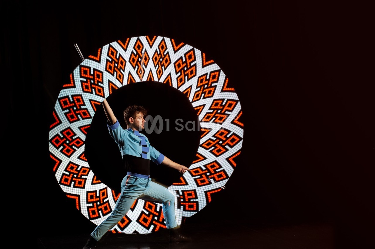 Un artiste en costume bleu exécute une pose dynamique devant un cercle lumineux à motifs colorés.