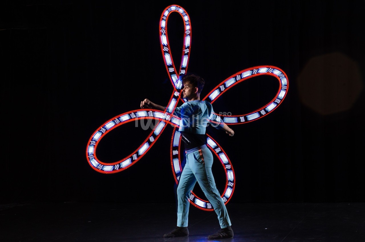 Un jongleur en costume bleu manipule des objets lumineux formant des motifs circulaires sur fond noir.
