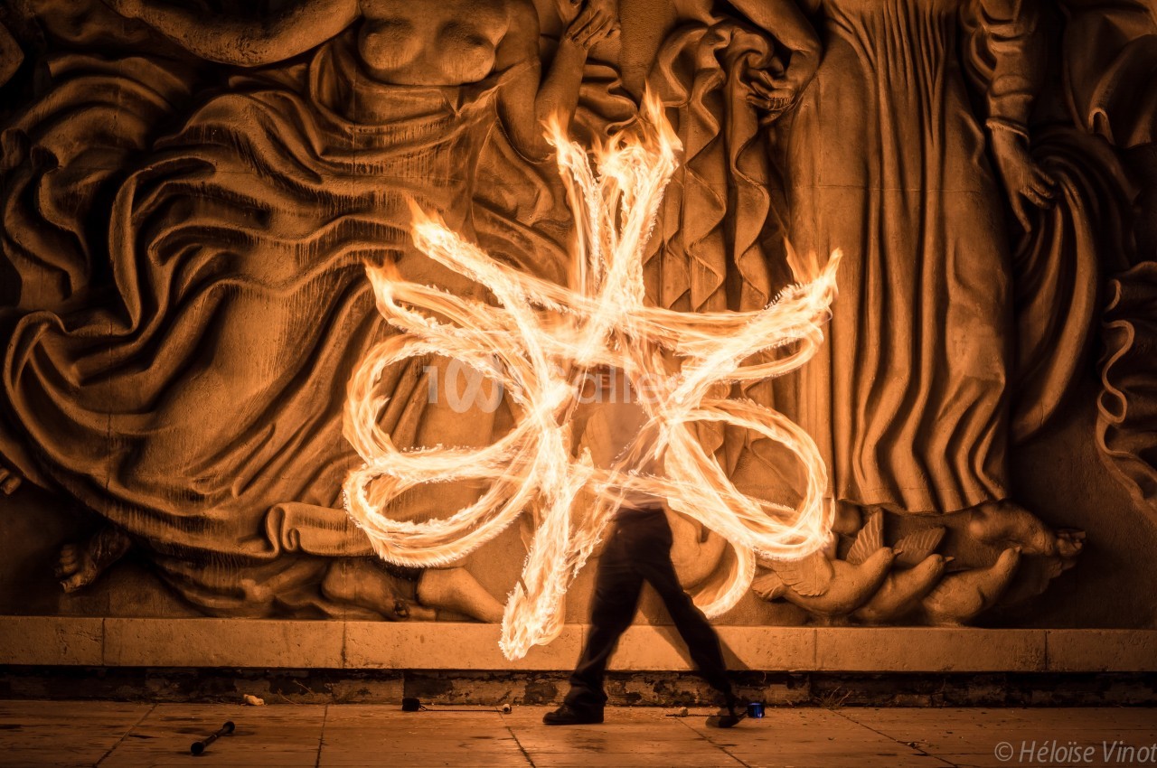 Personne jonglant avec des torches enflammées devant une sculpture murale éclairée dans un environnement nocturne.