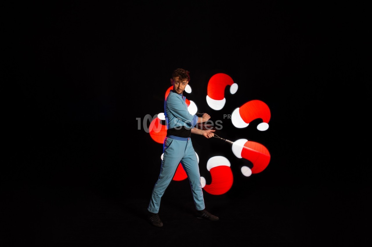 Un jongleur en tenue bleue manipule des bâtons lumineux créant des motifs rouges et blancs sur fond noir.