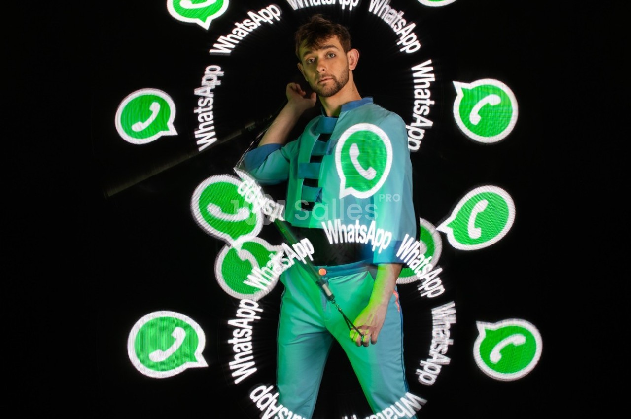 Un homme en tenue bleue pose devant un fond noir avec des logos WhatsApp lumineux flottant autour de lui.