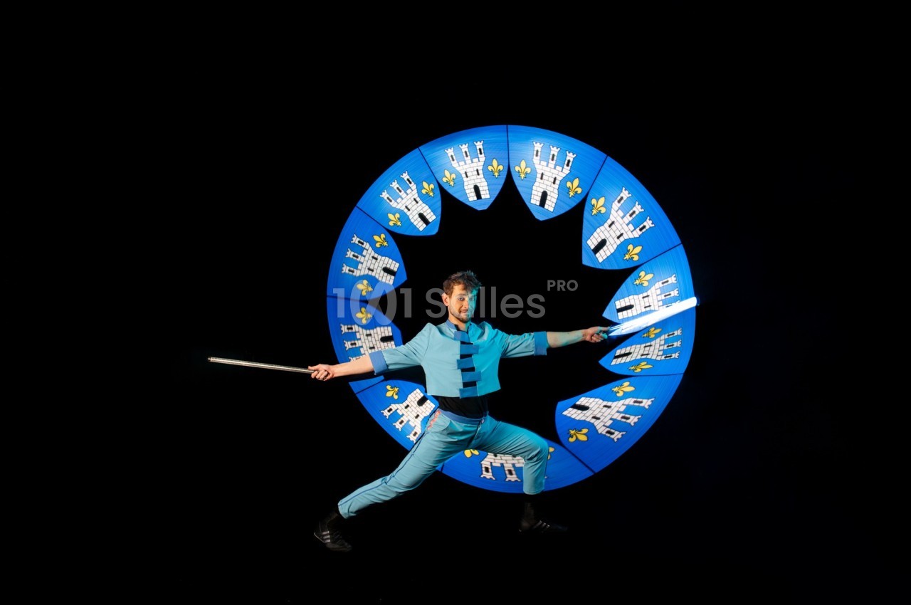 Un artiste en costume bleu manipule un bâton lumineux devant un cercle décoré de châteaux et de fleurs de lys.