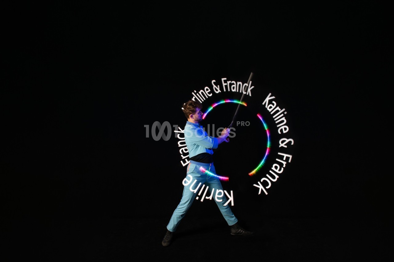 Un jongleur en tenue bleue manipule un bâton lumineux créant des motifs circulaires avec le texte ’Karine & Franck’.