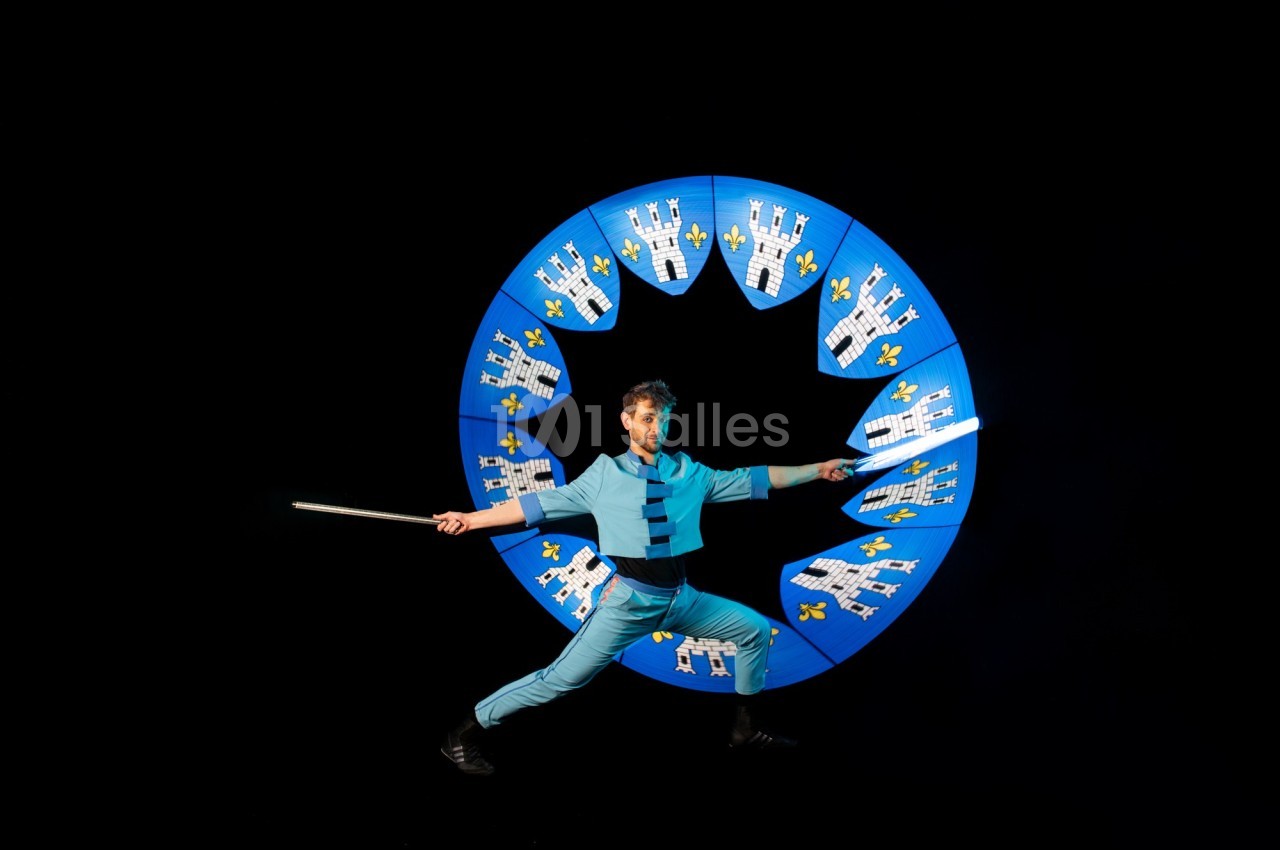 Un artiste en costume bleu manipule des bâtons lumineux devant un cercle décoré de châteaux et de fleurs de lys.