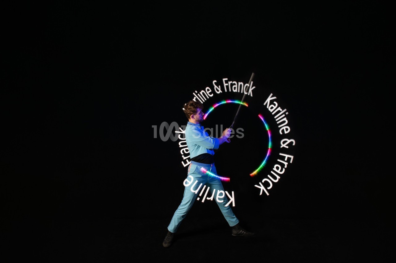 Un jongleur manipule un bâton lumineux créant des motifs circulaires colorés avec le texte ’Karine & Franck’.