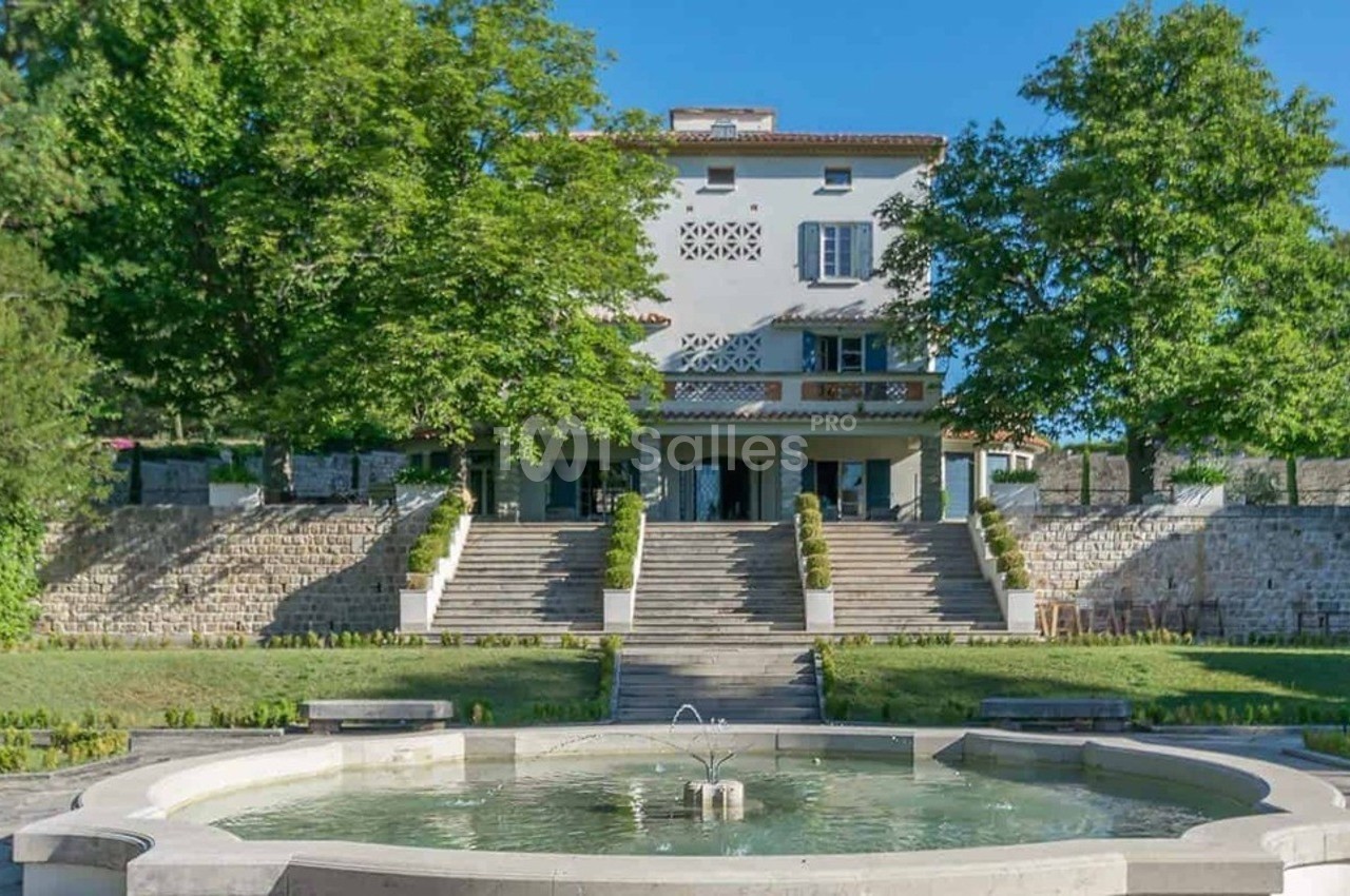 Grande maison blanche entourée d'arbres, avec un escalier menant à une fontaine circulaire au premier plan.
