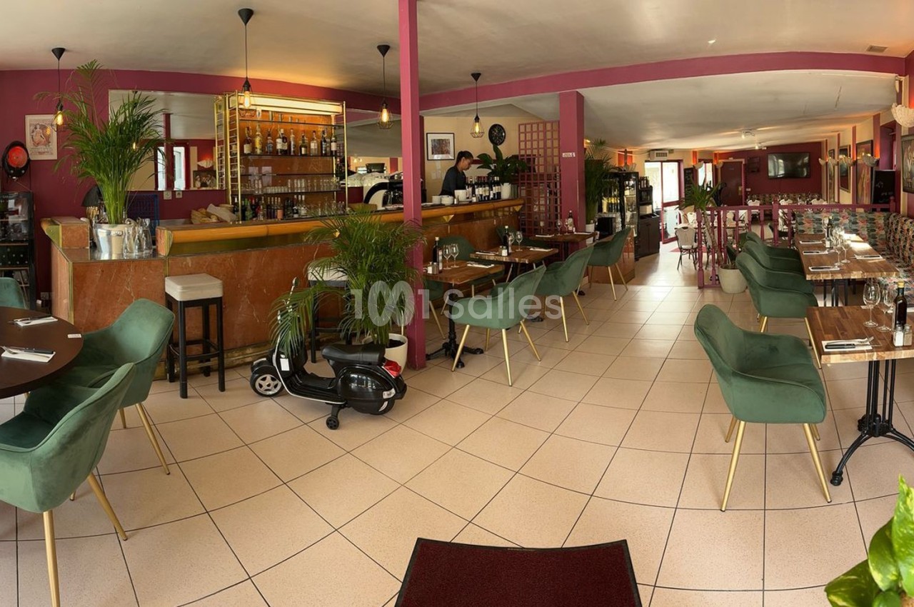 Salle de restaurant lumineuse avec tables en bois, chaises vertes, décorations végétales et comptoir en arrière-plan.