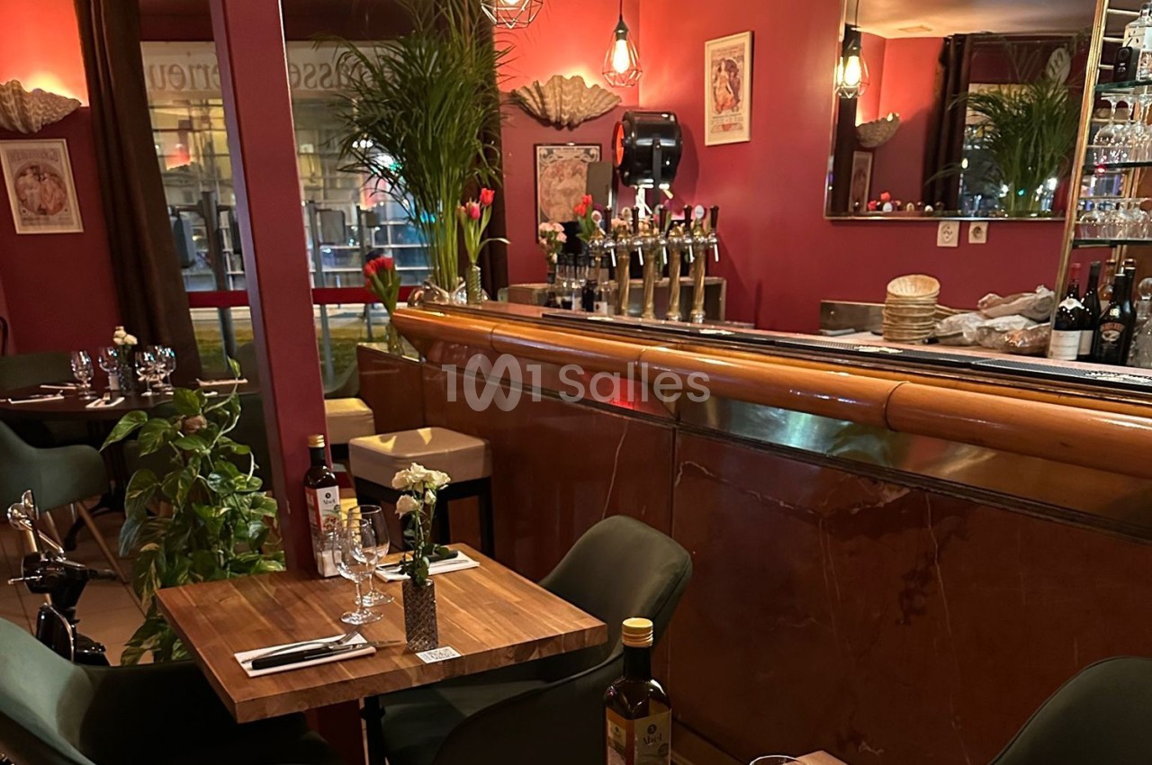 Salle de restaurant chaleureuse avec tables dressées, bar en bois, décor rouge et éclairage tamisé.