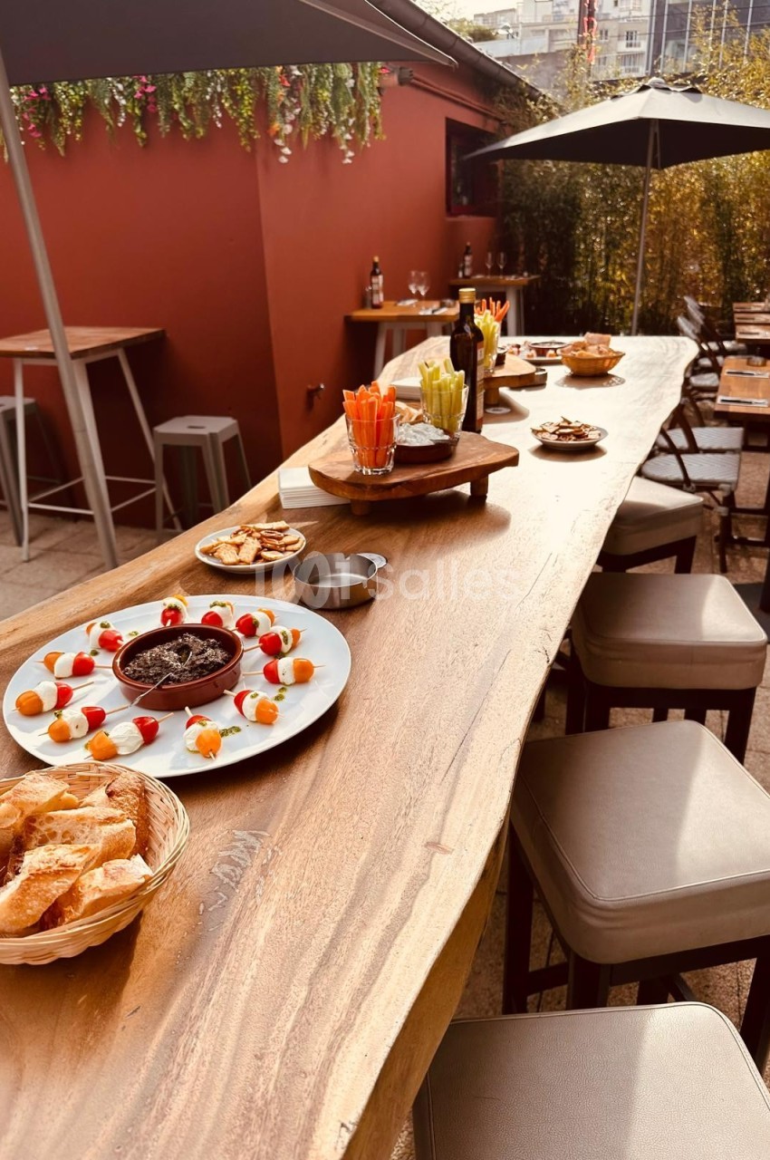 Table en bois avec apéritifs variés, légumes, dips et pain, disposée en terrasse avec chaises et parasols.
