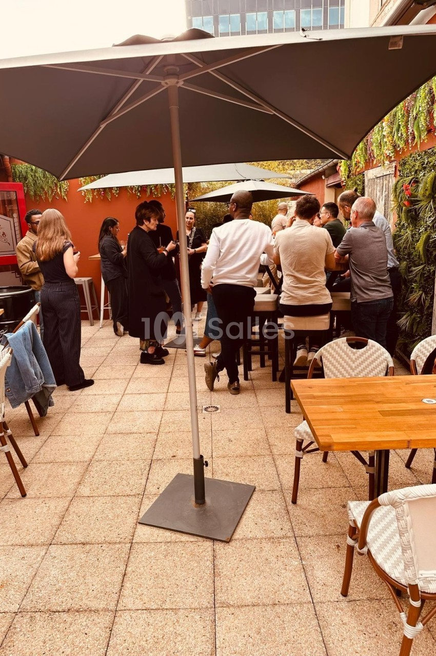 Personnes debout discutant sous un grand parasol sur une terrasse extérieure aménagée avec tables et chaises.