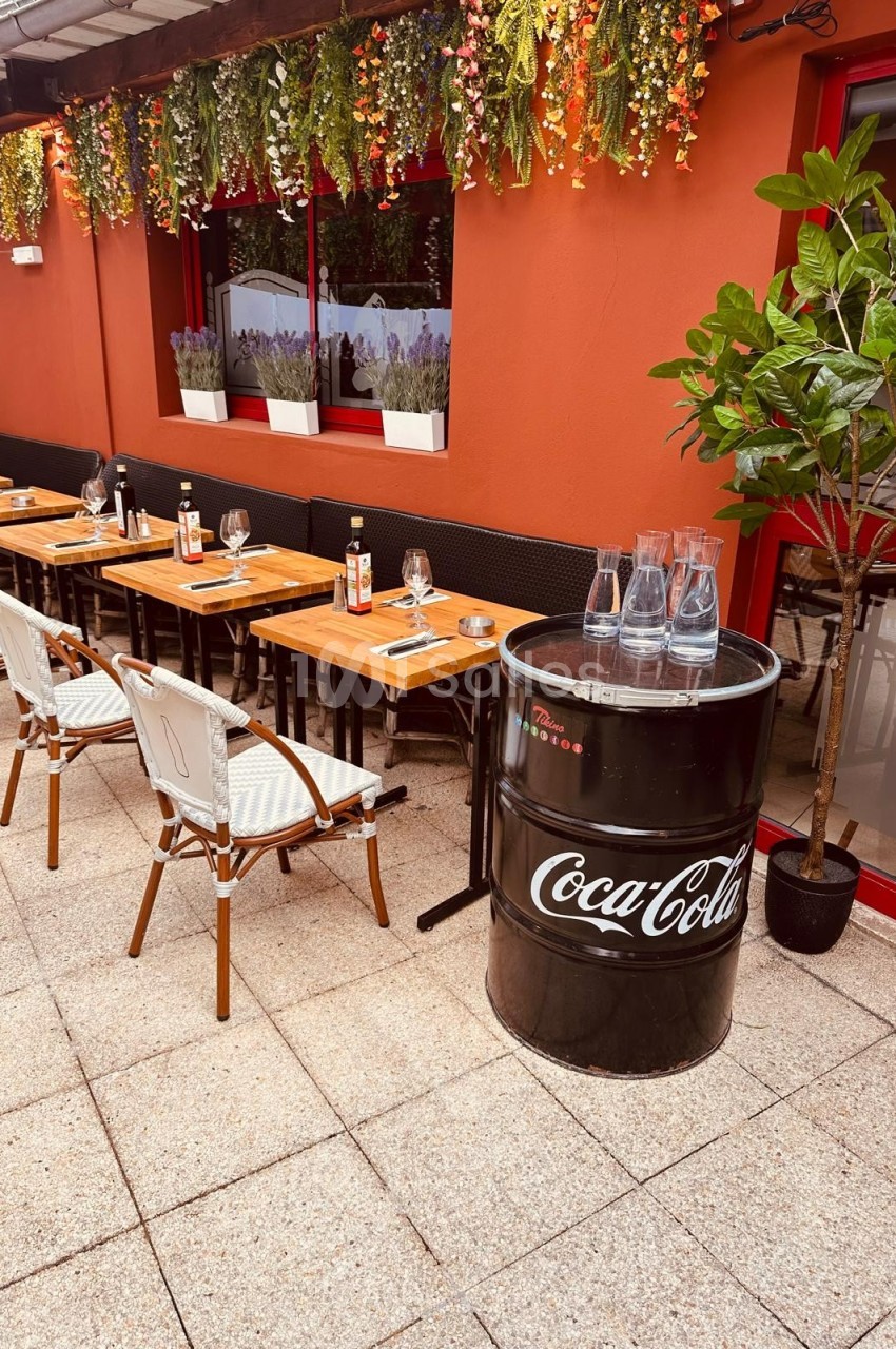 Terrasse d'un restaurant avec tables en bois, chaises blanches, plantes suspendues et un tonneau noir marqué Coca-Cola.