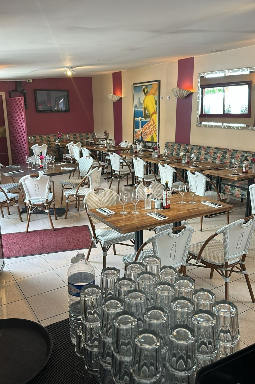 Salle de restaurant avec tables dressées, chaises en rotin, affiches murales et lumière naturelle entrant par une fenêtre.