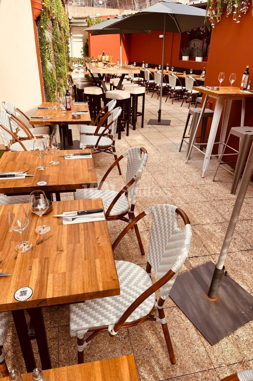 Terrasse d'un restaurant avec tables en bois, chaises tressées et parasols, dans un cadre extérieur ensoleillé.