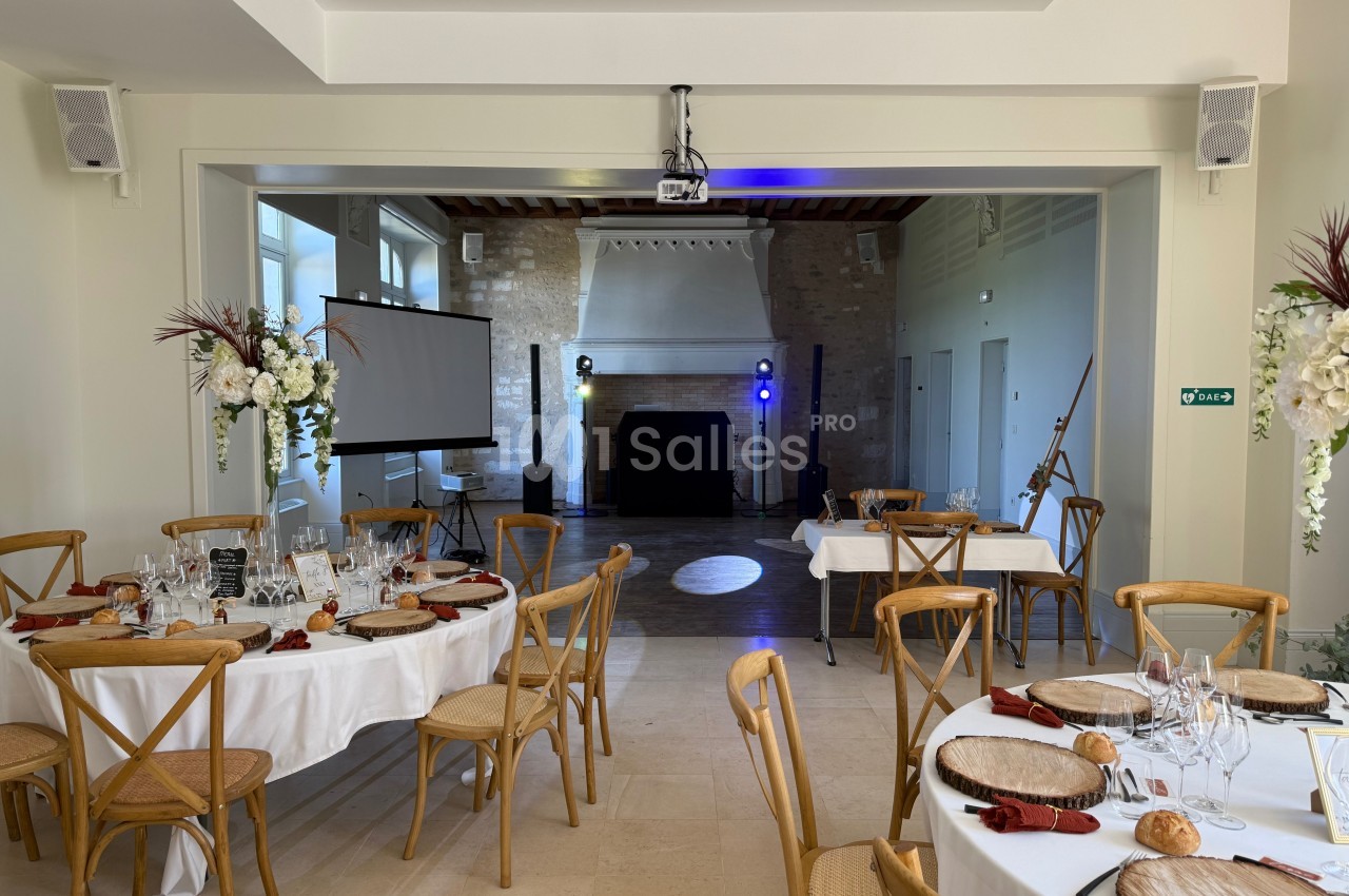 Salle de réception lumineuse avec tables dressées, écran de projection, projecteurs et décoration florale.