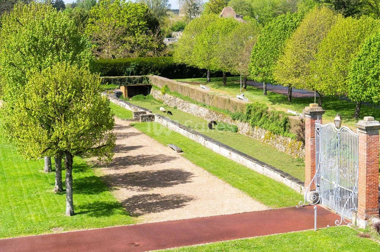 Allée bordée d'arbres et pelouses, avec un mur de pierre et une grille métallique dans un parc verdoyant.
