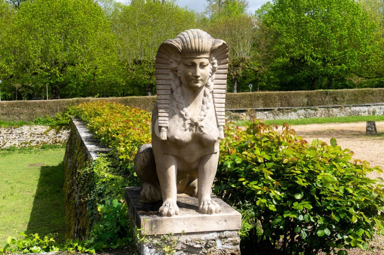 Statue de sphinx en pierre posée sur un socle, entourée de végétation et d'une haie dans un jardin.