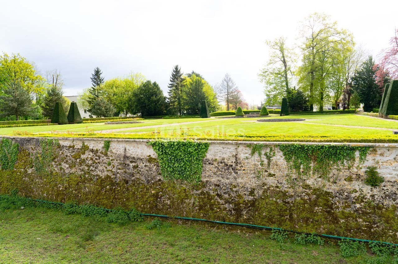 Mur en pierre recouvert de mousse et de lierre, surmonté d'un jardin verdoyant avec arbres et pelouses.