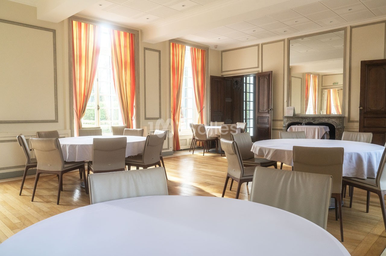 Salle lumineuse avec des tables rondes dressées de nappes blanches, entourées de chaises beige, et des rideaux orange.