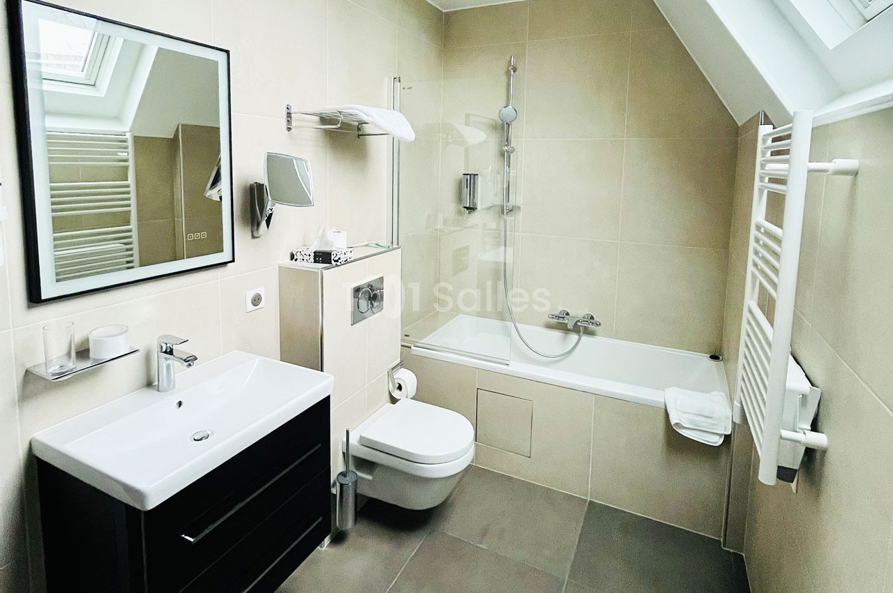 Salle de bain moderne avec baignoire, lavabo, toilette suspendue, miroir et radiateur mural sous une fenêtre de toit.