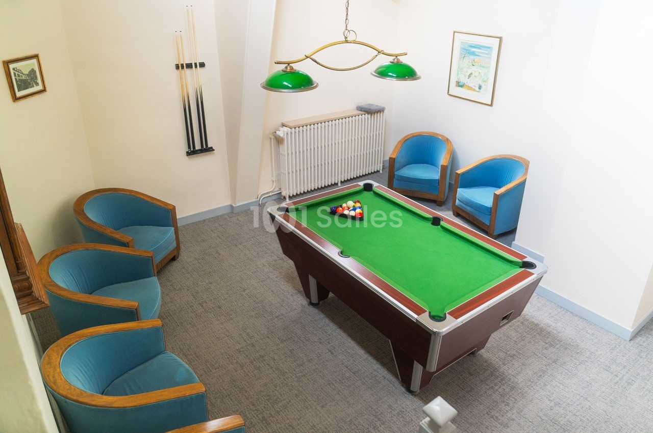 Salle avec une table de billard, quatre fauteuils bleus et des queues de billard accrochées au mur.