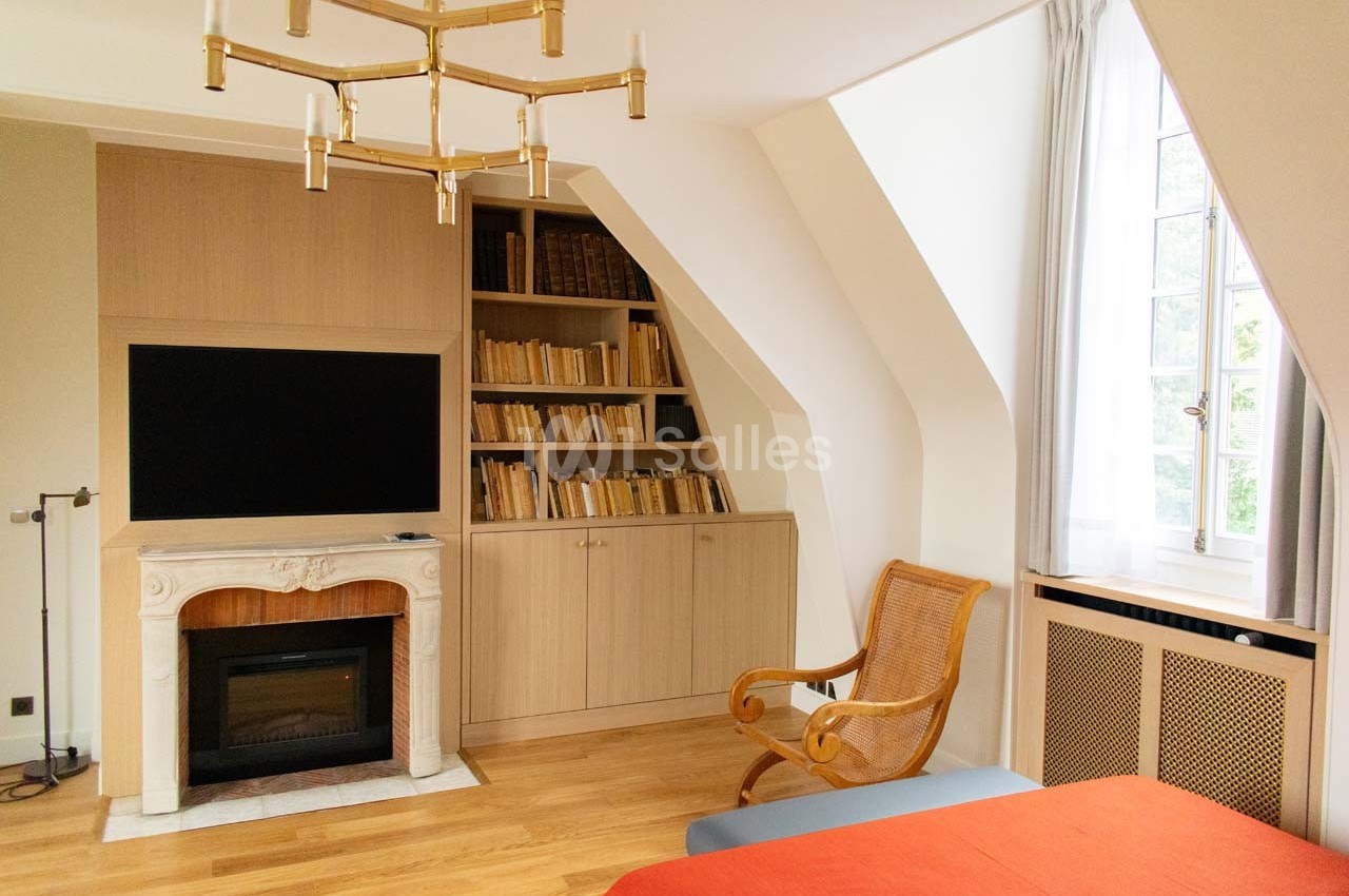 Chambre lumineuse avec parquet, cheminée décorative, étagères remplies de livres et fauteuil en osier près d'une fenêtre.