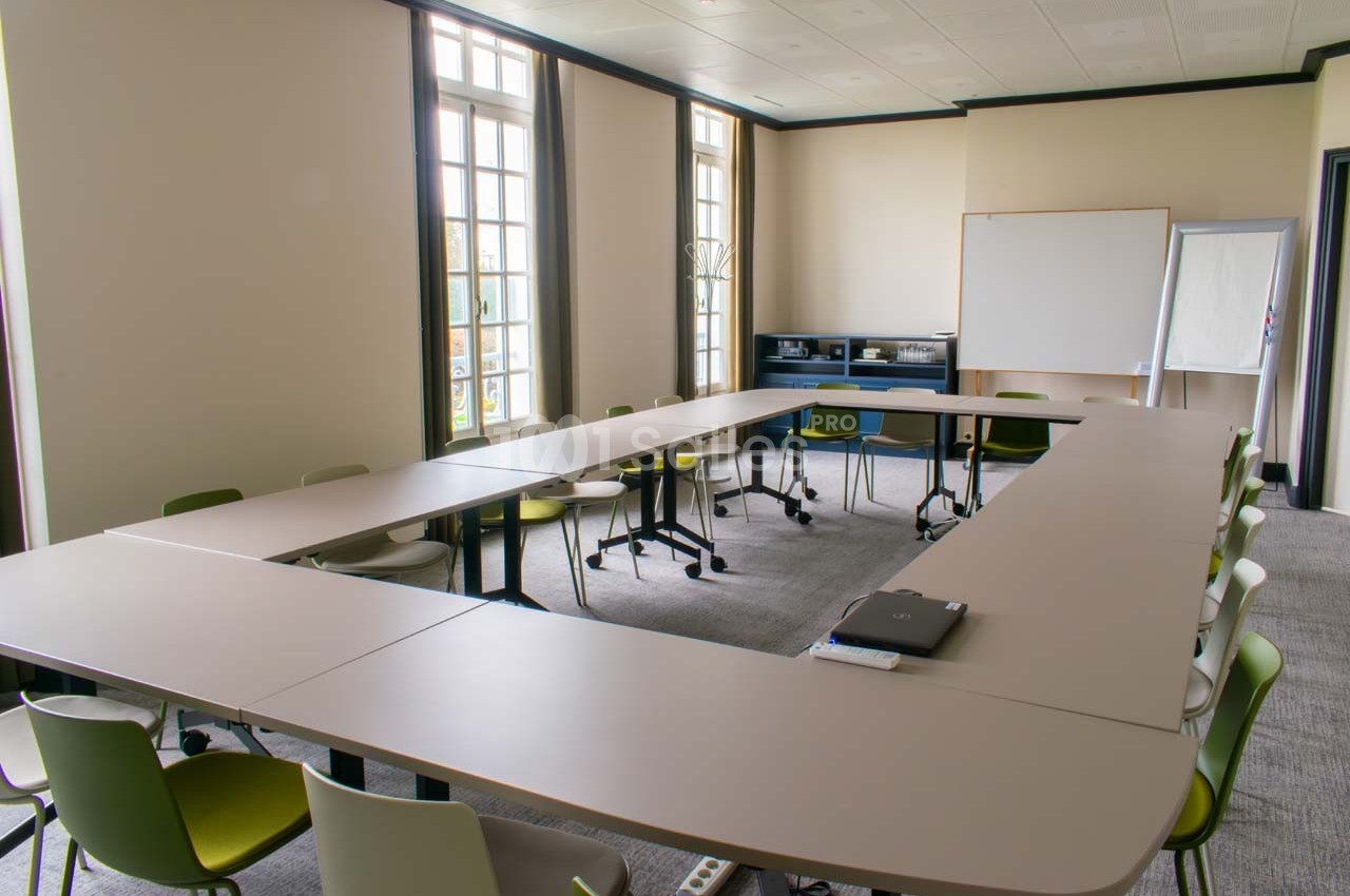 Salle de réunion lumineuse avec des tables disposées en U, des chaises colorées et un tableau blanc au fond.