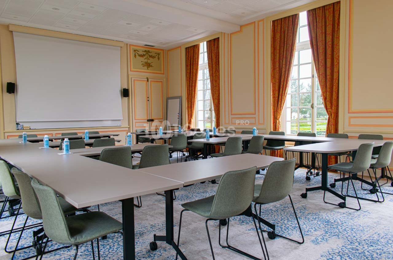 Salle de réunion lumineuse avec tables en U, chaises modernes, écran de projection et grandes fenêtres donnant sur un jardin.