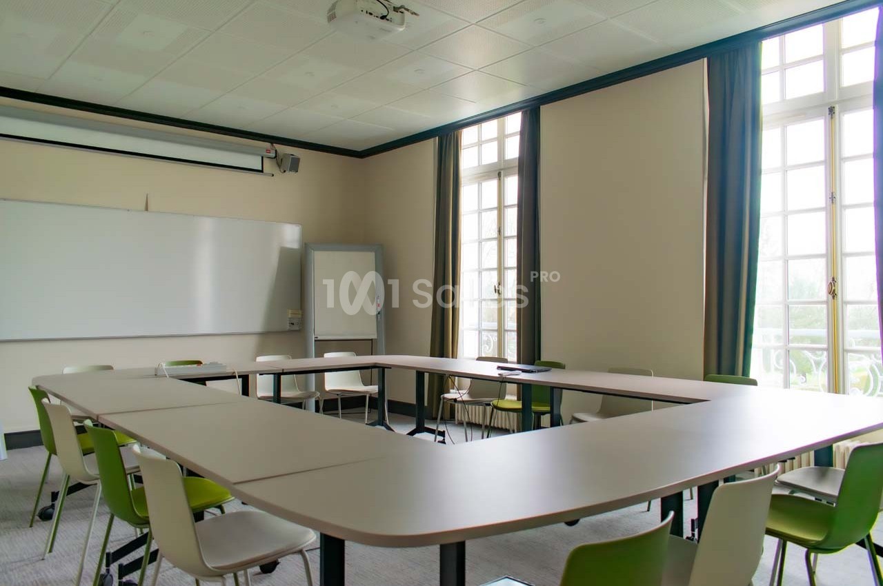 Salle de réunion lumineuse avec tables disposées en U, chaises vertes et blanches, tableau blanc et grandes fenêtres.