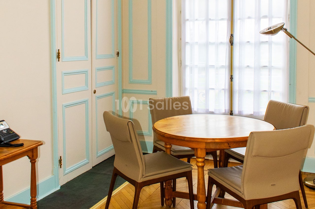 Salle lumineuse avec une table ronde en bois entourée de quatre chaises, près d'une fenêtre avec rideaux translucides.