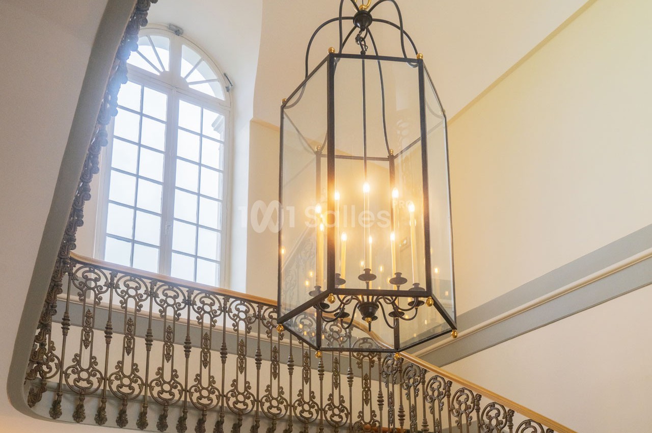 Lustre suspendu en métal noir avec bougies électriques, éclairant un escalier orné de balustrades en fer forgé.
