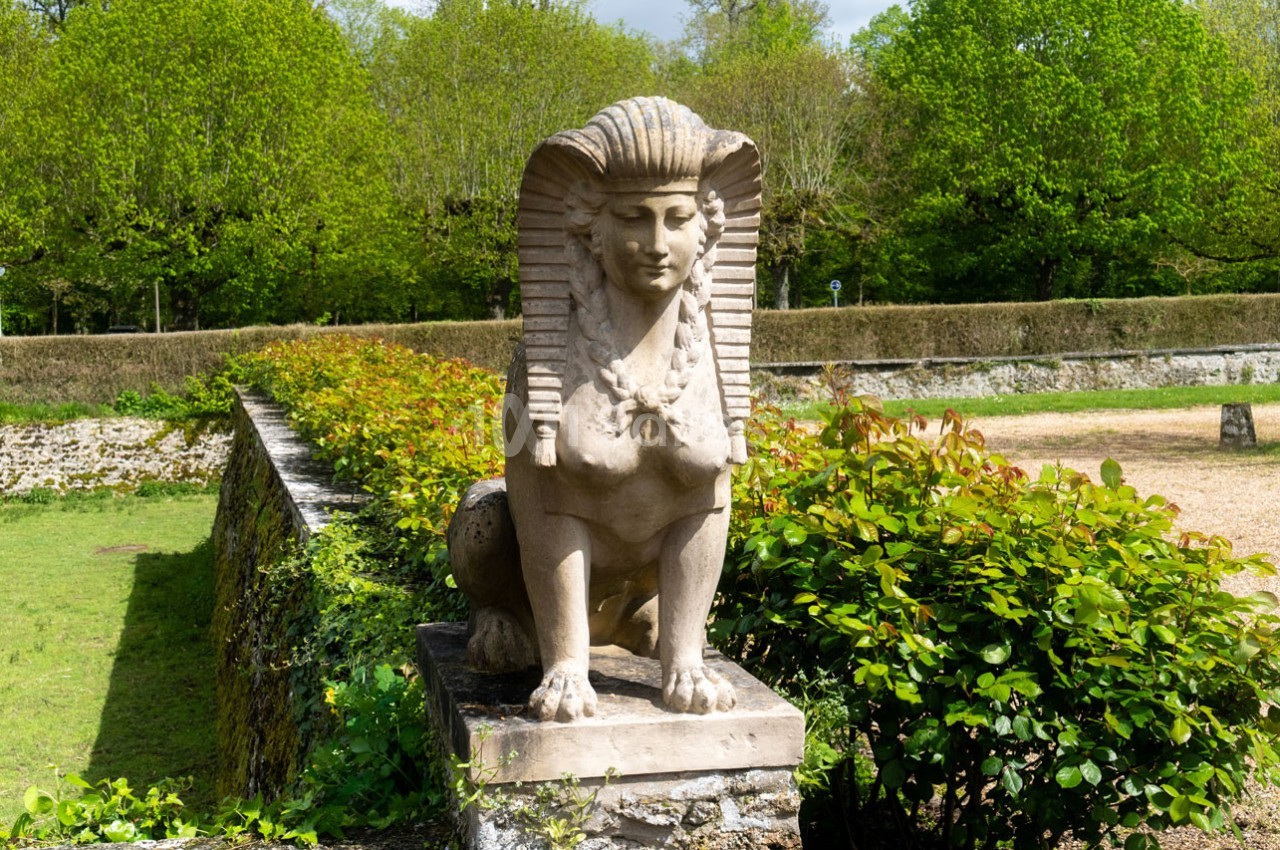 Statue de sphinx en pierre posée sur un socle, entourée de verdure dans un jardin.