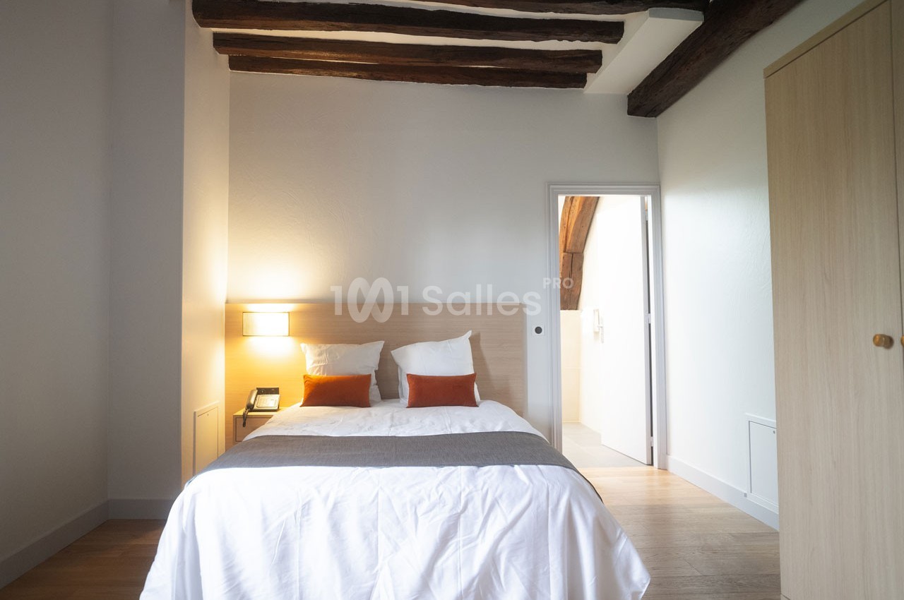 Chambre lumineuse avec lit double, poutres apparentes, armoire en bois clair et vue sur une salle adjacente.
