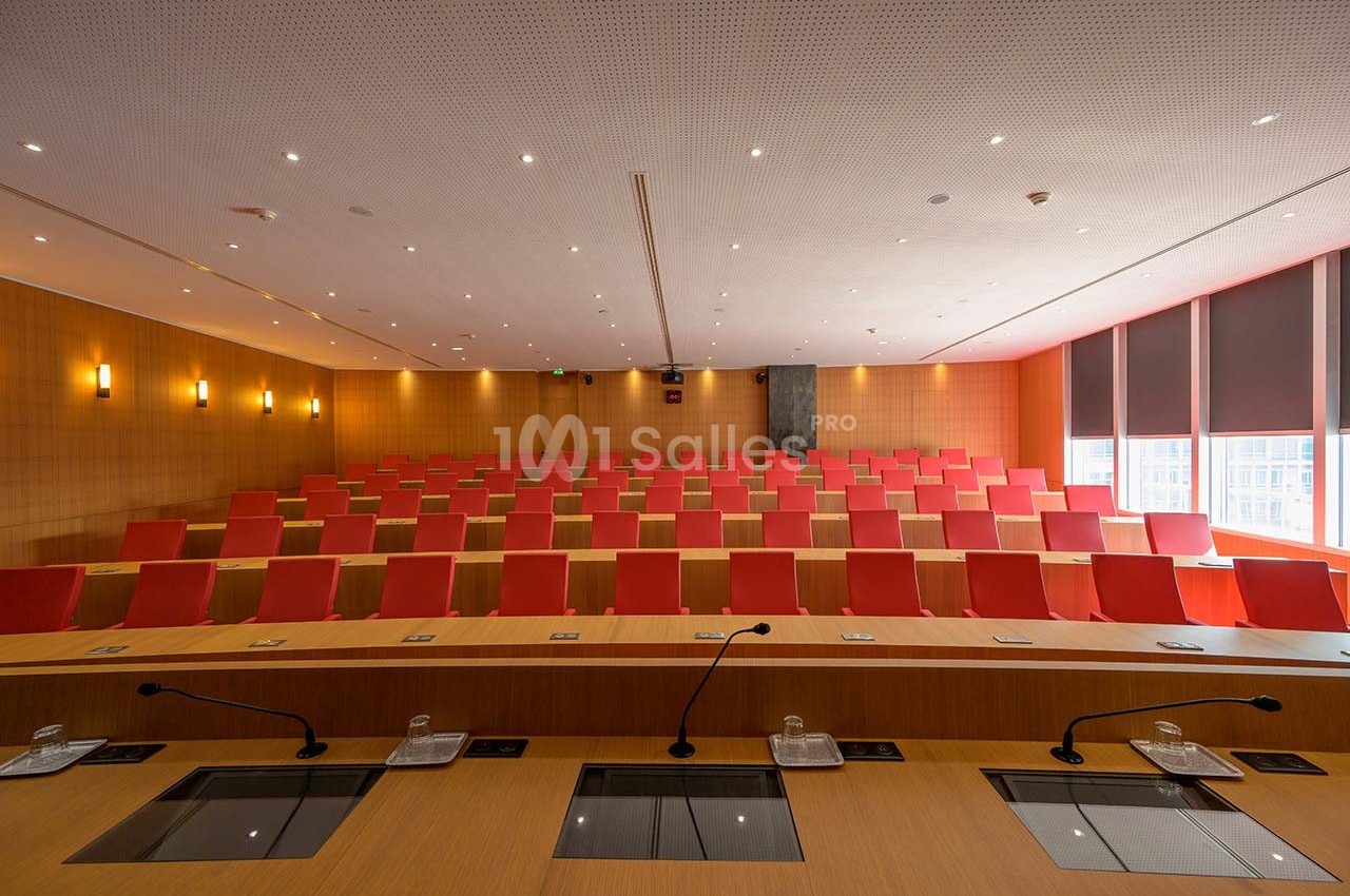 Salle de conférence vide avec rangées de sièges rouges, pupitres équipés de micros et éclairage tamisé.