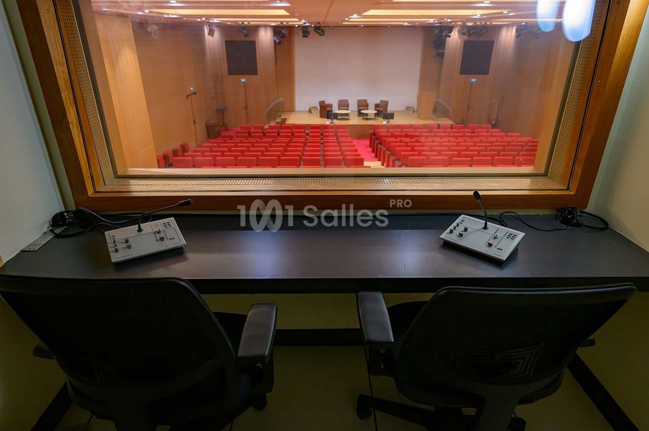 Vue depuis une cabine d'interprétation sur une salle de conférence avec des sièges rouges et une scène équipée de chaises.