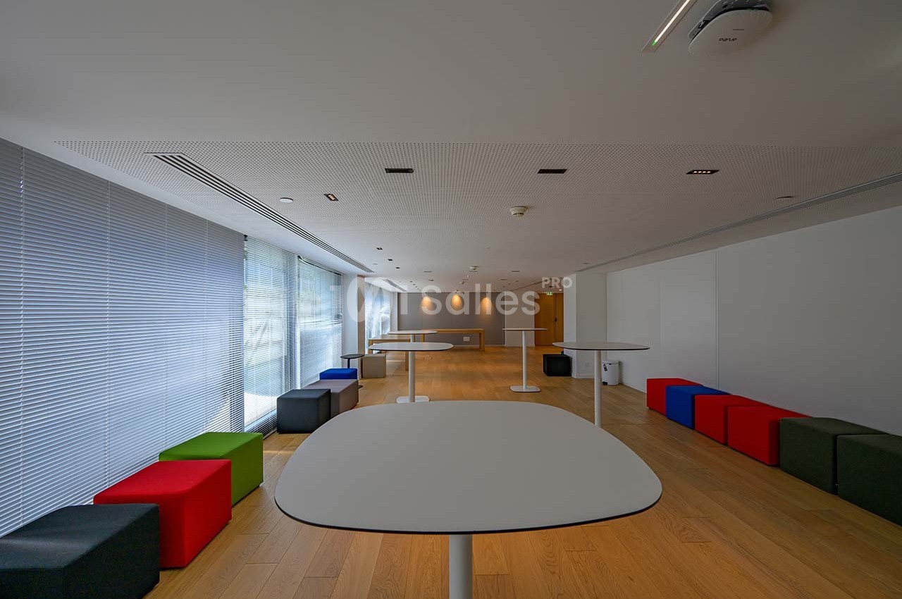Salle lumineuse avec tables blanches, tabourets colorés et parquet en bois clair, bordée de grandes fenêtres.